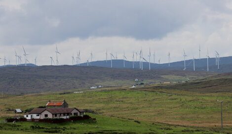 A rural landscape featuring a house and several farm buildings in the foreground, with a large wind farm on the hills in the background under a cloudy sky.