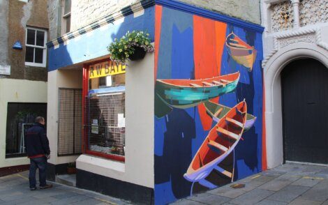A colorful mural of canoes on a building corner with a man looking at a window display, next to a flower basket hanging overhead.