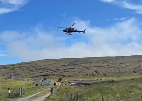A helicopter carrying materials via a cable over a construction site on a grassy hillside under a clear sky. workers and machinery are visible on the ground.