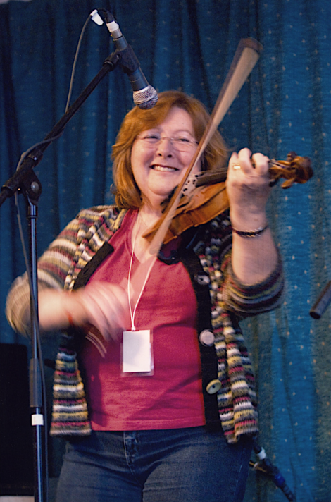 Woman playing violin enthusiastically on stage, smiling, with a microphone above her and a blue draped background.