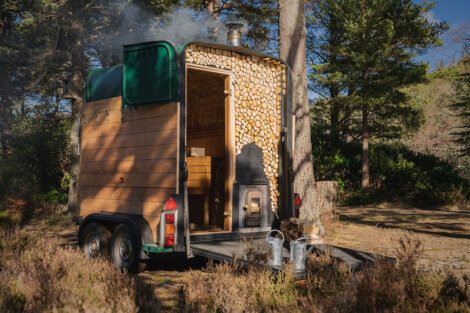 Mobile wooden sauna with a smoking chimney parked in a forest clearing.
