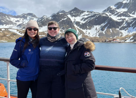 Three individuals smiling for a photo on a ship with a snowy mountainous backdrop.