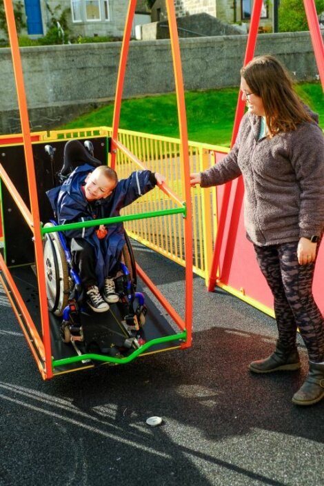 A woman assists a child in a wheelchair on an accessible swing at a playground.