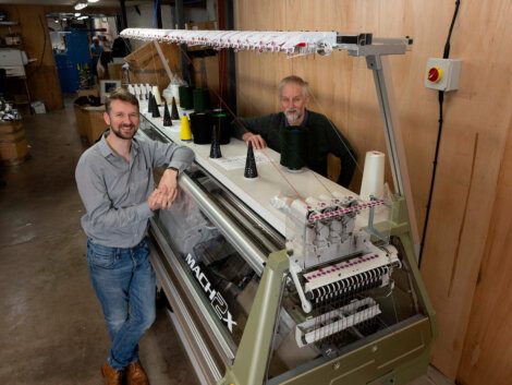 Two men smiling at the camera while standing beside an industrial embroidery machine in a workshop setting.