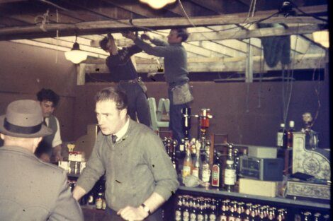 Men working on electrical wiring above a bar while a patron and bartender carry on below.