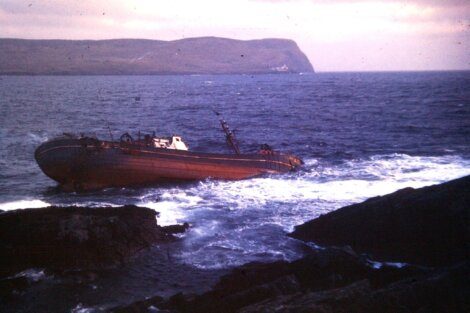 Shipwrecked vessel lying on its side near rocky coastline with waves crashing around.