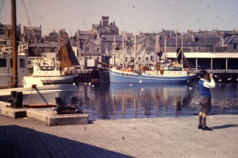 A person taking a photograph at a harbor with moored fishing boats and buildings in the background.