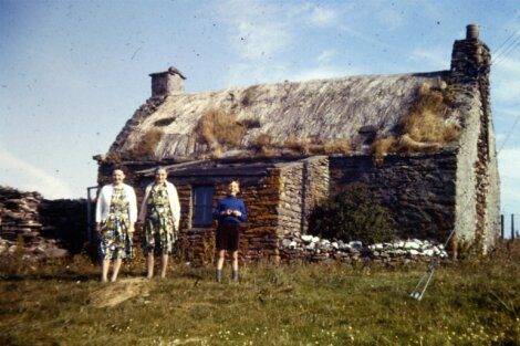 Three individuals standing in front of a traditional thatched-roof cottage on a sunny day.