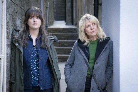 Two women standing in an alleyway, looking away from the camera with curious expressions.