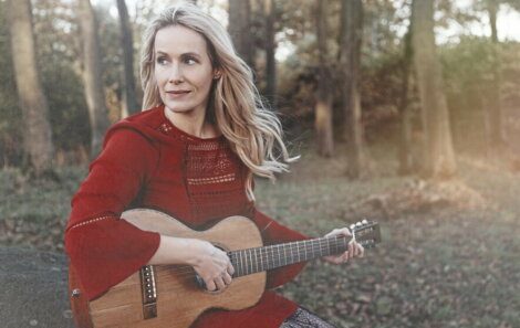 A woman is sitting in the woods with an acoustic guitar.