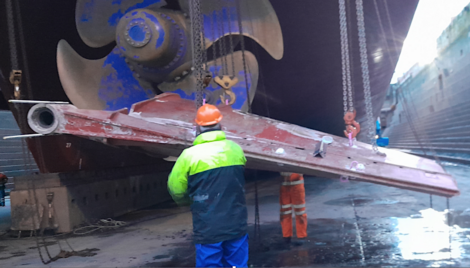 A worker in a high-visibility jacket observes a large ship's propeller being hoisted by a crane in a dry dock facility.