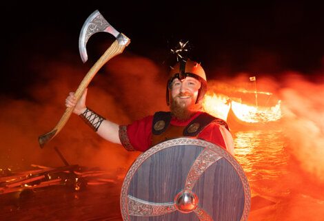 Person dressed as a viking holding an axe with a burning boat in the background.