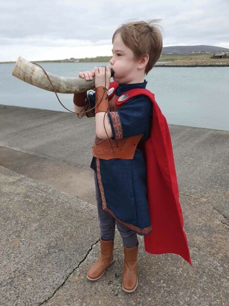 Child dressed in a viking costume blowing into a horn by the waterside.