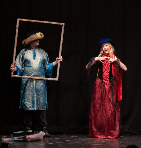 Two actors on stage, one holding a frame as if to be a portrait, the other wearing a cap and gown performing a gesture.