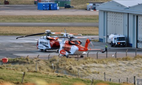 Two helicopters on a tarmac near an airport building with ground support equipment in the vicinity.