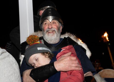 A bearded man in medieval armor smiling and holding a sleeping child at a nighttime event with torches in the background.