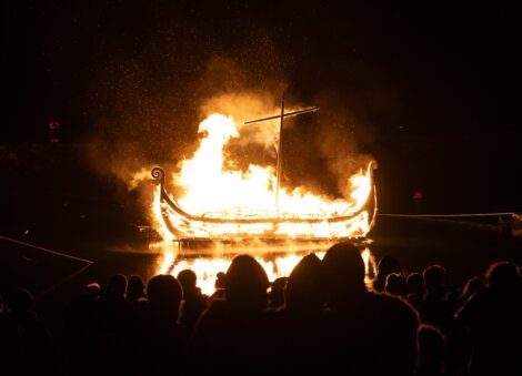 A silhouette of spectators watching a viking ship effigy ablaze during a night-time ceremony.