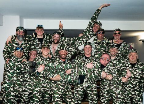 A group of people wearing matching camouflage outfits and various hats, posing for a photo with smiles and playful gestures.