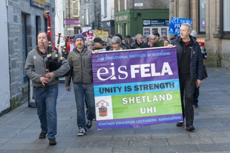 A group of demonstrators marching on a street, led by a bagpiper, while carrying a banner for the further education lecturers' association advocating for unity and strength.