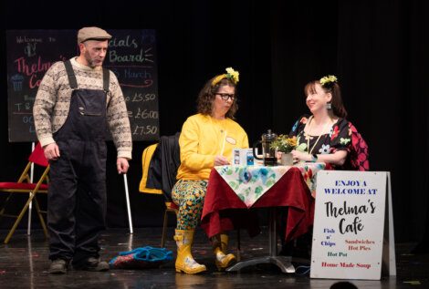 Three actors on stage in a scene set in a cafe, with one standing and two seated at a table, engaging in conversation amidst props and a menu sign.