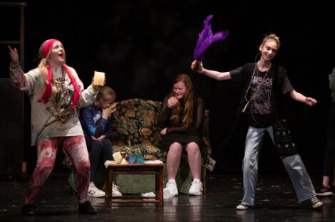 Two individuals performing on stage with expressive gestures, one holding a feather, while others sit on a couch, one covering her face and laughing.