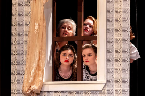 Four women of varying ages peer through a window frame on stage, suggesting a scene from a theatrical performance.