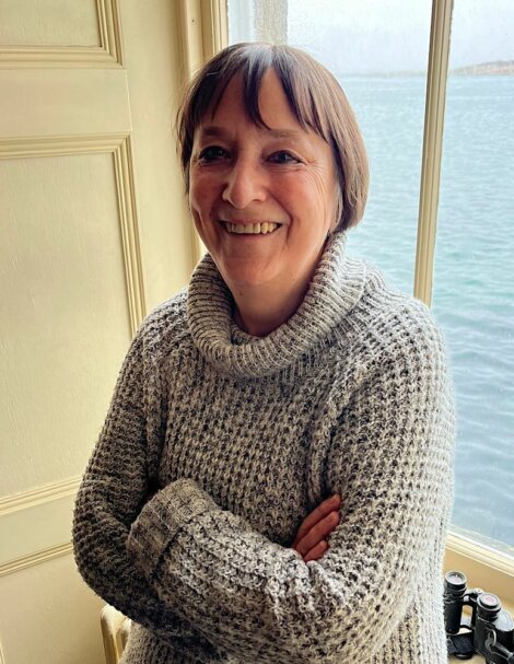 A smiling middle-aged woman in a knitted sweater standing by a window with a view of the sea.