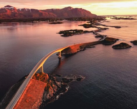 Aerial view of a curvy bridge crossing over a series of small islands in a calm sea at sunset, with mountains in the background.