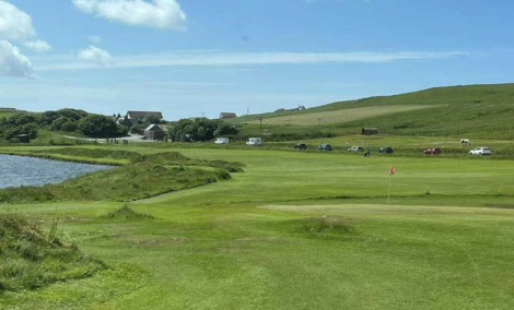 A scenic golf course with lush green grass, a small pond on the left, few cars parked in the distance under a clear blue sky.