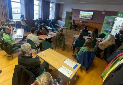 A group of people seated in a conference room, attentively watching a presentation on a screen, with nature posters on the walls.