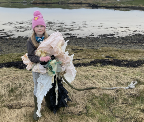 Young girl holding large pieces of plastic waste by a lakeside, wearing a pink hat and a brown jacket, promoting environmental cleanup.