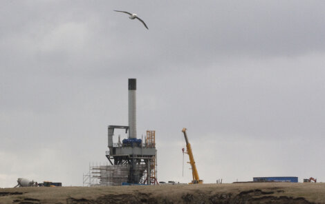A seagull flies over an industrial construction site with large machinery, cranes, and a tall chimney under a cloudy sky.