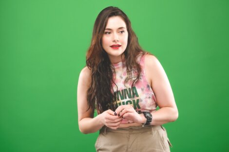 A young woman wearing a tie-dye tank top and khaki pants stands against a green backdrop, looking surprised as she checks her watch.